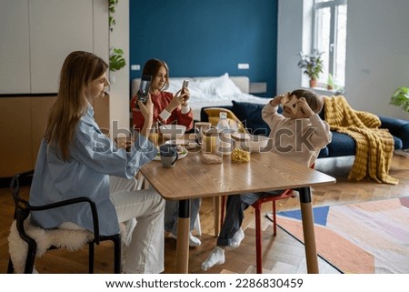 Family breakfast with addiction from smartphone. Pleased mom takes pictures as kid son fooling around with bread toast while daughter look at screen cellphone at kitchen table in morning before school