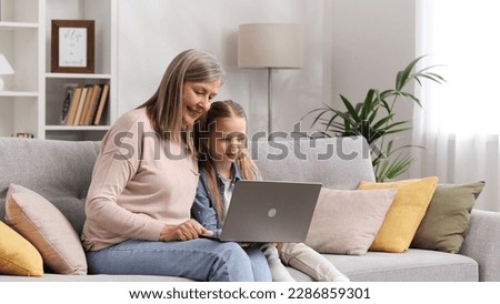 Grayhaired grandmother and little granddaughter are sitting on the sofa in the living room, resting and watching funny video cartoons on the laptop, talking, having fun.