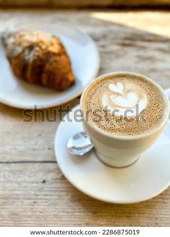 Cup of fresh flat white or latte coffee with croissant on wooden background. French style breakfast in a cafe. Latte art coffee with freshly baked croissant on a wooden table. pov.