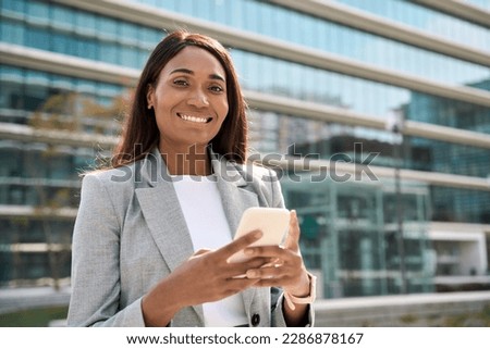 Portrait of young beautiful successful businesswoman, African American business woman, sales leader looking at camera, holding mobile phone application, using cellphone in hand outdoor office center. 
