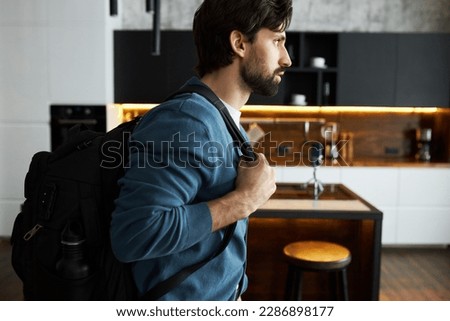 Side view closeup of handsome bearded business man in blue cardigan carrying black backpack on shoulder leaving home, going to adventurous trip, pictured against kitchen background