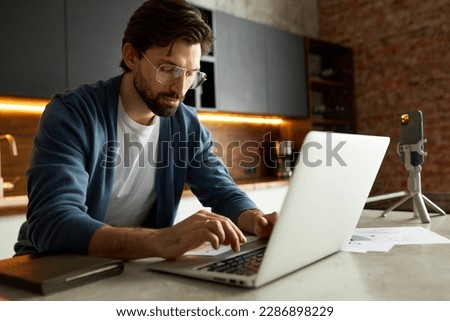 Intelligent smart guy with beard in stylish glasses sitting at kitchen table working on laptop recording video for podcast, typing on keyboard, preparing for live stream. Content creator studying