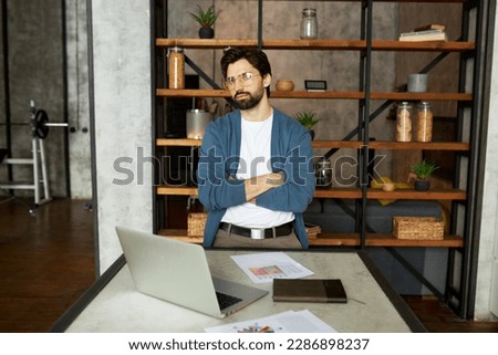 Bossy successful financial expert or trader standing next to working table with laptop and paper documents with crossed hands in loft style home interior, satisfied with his new achievements Royalty-Free Stock Photo #2286898237