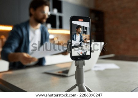 Selective focus on tripod with smartphone recording male business coach sitting at kitchen table in front of laptop, holding paper documents, preparing for live stream. Vlogging concept Royalty-Free Stock Photo #2286898247