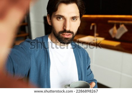 Image of attractive hipster guy with bearded face making selfie on kitchen background, holding cup of coffee in hands, spending leisure time, having early morning breakfast before going to work