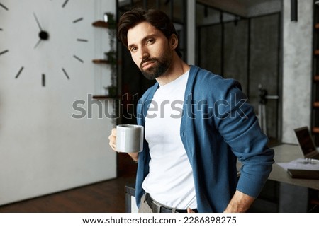 Charming male head of customer support standing at his office with cup of coffee or tea, looking at camera, wearing blue cardigan upon white mockup t-shirt, pictured against big wall clock