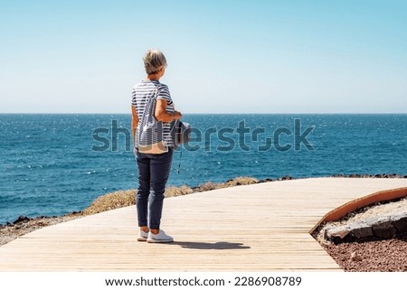 Senior woman standing in front to the sea in summer holidays looking at the horizon. White haired lady dressed in blue in vacation or retirement Royalty-Free Stock Photo #2286908789