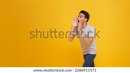 Young Asian man shouting open mouths raising hands screaming announcement to empty space aside isolated on yellow studio background. Royalty-Free Stock Photo #2286911571