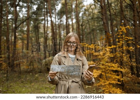 Beautiful blonde woman holding mobile phone using map to navigate in autumn forest. Happy tourist trying to find best way. Active lifestyle, travel concept. High quality photo