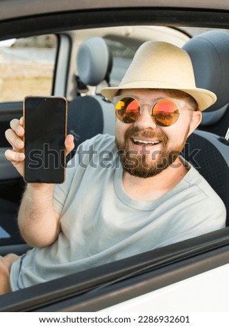 Smiling bearded man in sunglasses showing mobile phone with empty screen for mock up sitting on driver seat in new luxury car cabriolet, holding gadget in hand, selective focus on device