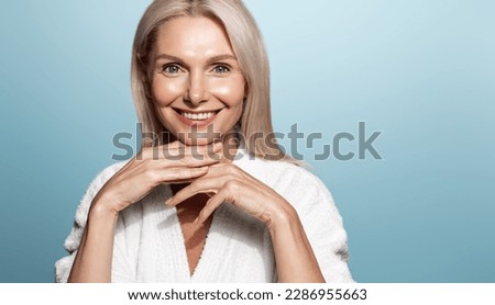 Smiling middle aged female model with wrinkles, nourished, glowing facial skin, moisturized face, looking happy at camera, blue background.
