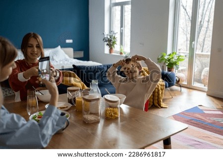 Pleased mother and teen girl take pictures on smartphone as kid boy fool around with bread toast at kitchen table in morning before school. Happy child son make eyes with food on family fun breakfast 
