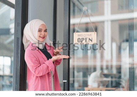 Portrait of a smiling young beautiful Muslim female owner of a startup coffee shop or restaurant turning the Open sign on the door