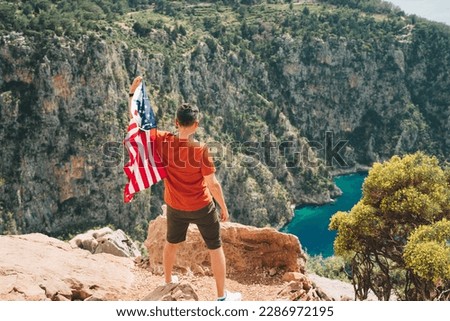 Young man standing on a rock cliff and waving the US flag while looking at sea beneath. Veteran traveller waving the American flag while standing on a mountain top. 4 fourth July Independence Day
