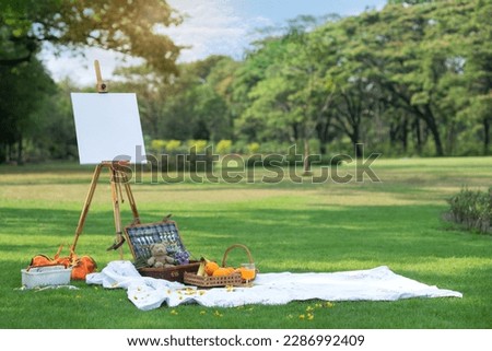 Picnic basket, fruits basket on picnic cloth, drawing and easel with frame on lawn outside in summer park, summer picnic background concept Royalty-Free Stock Photo #2286992409