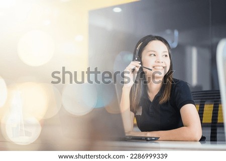 Friendly Asian female officer smiling happily in headset working on computer in call center.