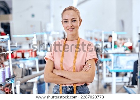 positive woman dressmaker standing in textile factory at workshop, attractive tailor after work stand posing, looking at camera smiling. female use measuring tape for sewing and making garments