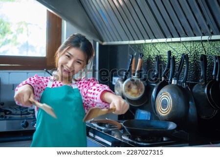 Beautiful asian cook wearing apron holding a paddle with both hands in kitchen and look at the camera with smile on her face.