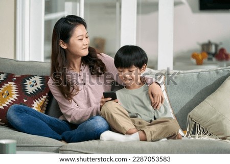 happy asian mother and son sitting on family couch looking at cellphone photos together