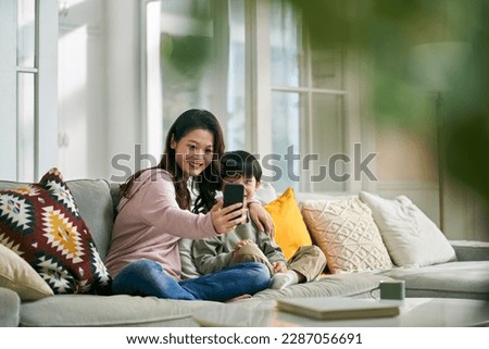 happy asian mother and son sitting on family couch at home taking a selfie using mobile phone