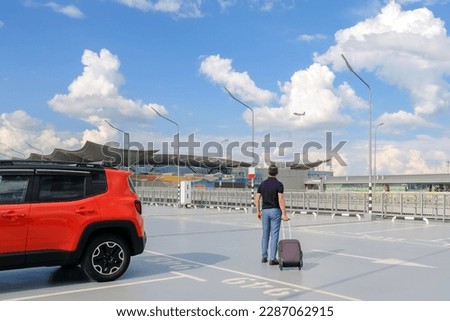 A young man with a suitcase in the airport parking lot sees the plane off with a look.