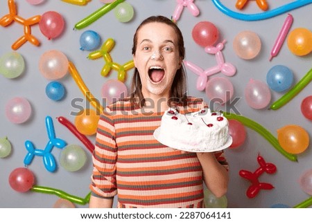 Happy joyful cheerful woman with brown hair wearing striped dress holding cake standing against gray wall decorated with colorful balloons posing with sweet dessert on her face, screaming with joy.