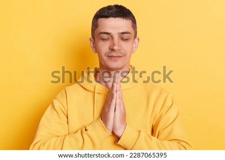 Meditation. Practicing yoga. Calm relaxed man wearing casual hoodie keeps palms together closed eyes doing yoga for relaxing posing isolated over yellow background.