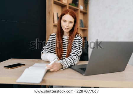 Portrait of positive young woman with broken right hand wrapped in white gypsum bandage working remotely with paper documents and laptop computer, smiling looking at camera, sitting at table.