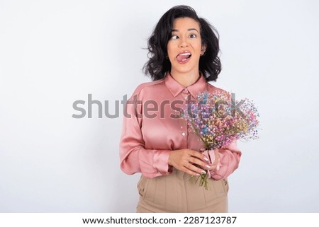 beautiful woman holding a bouquet of flowers over white background  showing grimace face crossing eyes and showing tongue. Being funny 