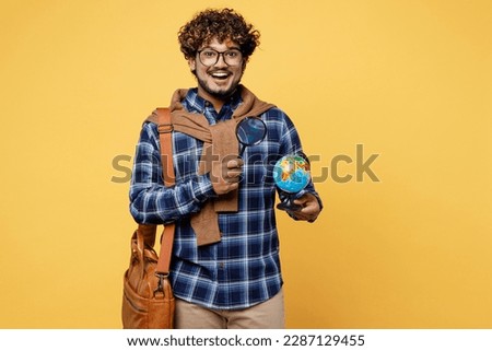 Young amazed teen Indian boy IT student he wear casual clothes glasses bag hold globe Earth map use magnifying glass isolated on plain yellow color background High school university college concept