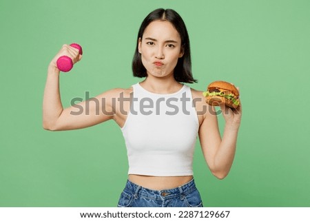 Young woman wear white clothes hold eat burger dumbbel burns calories do sport workout show muscles isolated on plain pastel light green background. Proper nutrition fast food unhealthy choice concept Royalty-Free Stock Photo #2287129667