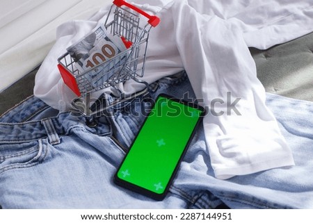 Shopping cart with American dollars, a smartphone with green screen, and different clothes. Concept of traveling or shopping