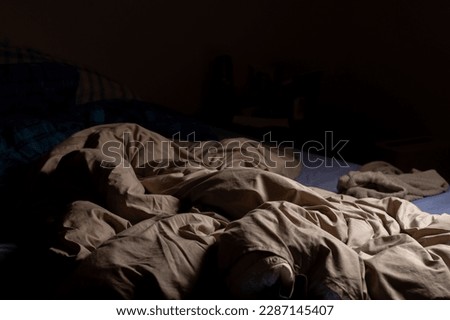 An empty unmade bed in the dark. A crumpled blanket lit by a lamp. Royalty-Free Stock Photo #2287145407