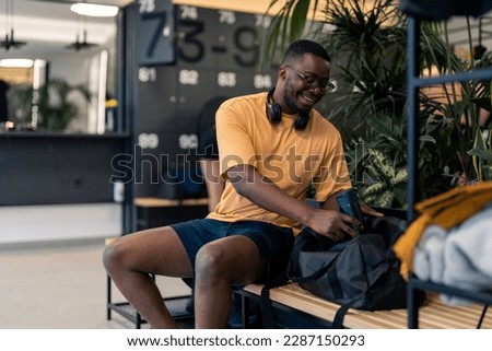 Handsome athletic man with headphones looking satisfied sitting on bench in locker room at gym, packing water bottle in sports gym bag after finished workout session, preparing to go home.