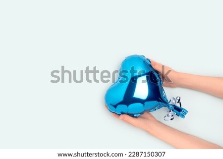 Female hands hold inflatable foil balloon in a heart shape. Minimal concept with place for text on a blue background.