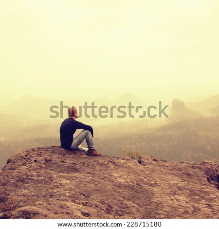 Tourist sit on peak of sandstone rock and watching into colorful mist and fog in  morning valley. Sad man. Man sit. Man in jeans.Fall mountain mist. Mist in valleys. Man hand.Sad tourist sit in mist. Royalty-Free Stock Photo #228715180