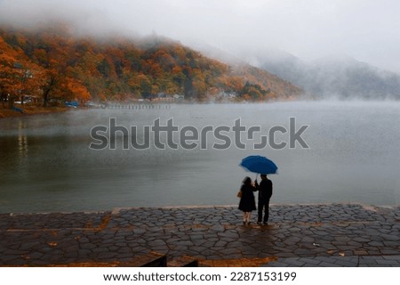 A couple stand under an umbrella on the paved promenade by Lake Chuzenji to enjoy the view of colorful foliage on the lakeside hills on a foggy rainy autumn day, in Nikko National Park, Tochigi, Japan