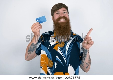 Smiling Red haired man wearing printed shirt over white studio background showing debit card pointing finger empty space