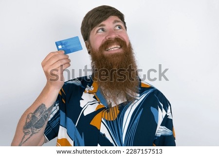 Lovely Red haired man wearing printed shirt over white studio background showing credit card and looking away at copy space. Pensive