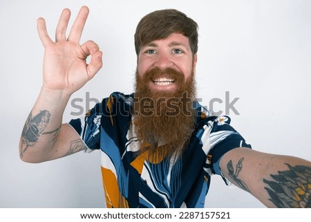 Portrait of pretty cheerful Red haired man wearing printed shirt over white studio background make selfie show okey symbol
