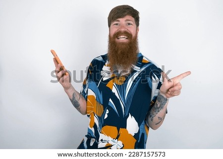Astonished Red haired man wearing printed shirt over white studio background holding her telephone and pointing with finger aside at empty copy space