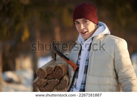 Active recreation in winter, travel and tourism. A young man in warm winter clothes holds an armful of firewood, standing in a winter snowy forest.