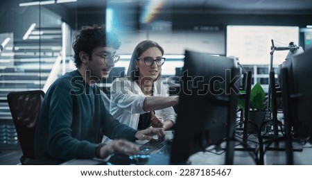 Two Young Colleagues Working on Computers and Talking at a Workplace. Female and Male Software Developers Discussing a Solution for Their Collaborative Artificial Intelligence Project Royalty-Free Stock Photo #2287185467