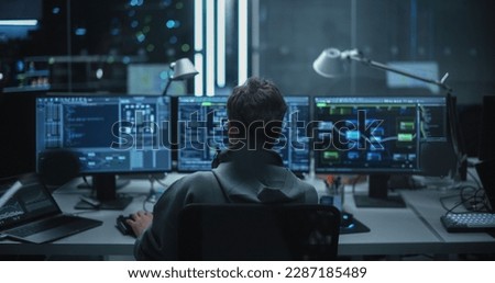 Lone Hacker Breaks Into Government Data Servers and Infects Their System with an Exploit Software. His Hideout Place is in Dark Technological Facility with Multiple Displays. Shot from the Back Royalty-Free Stock Photo #2287185489