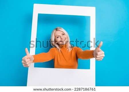 Portrait of optimistic friendly woman blond hairstyle dressed orange sweatshirt show thumb up from frame isolated on blue color background