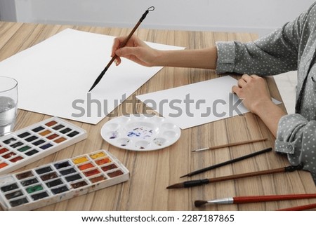 Woman painting with watercolor on blank paper at wooden table, closeup