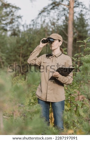 A woman park ranger in uniform looks through binoculars at the forest area with a clipboard in hand in summer, vertical photo, selective focus. Ecologist, national park, forester, nature concept
