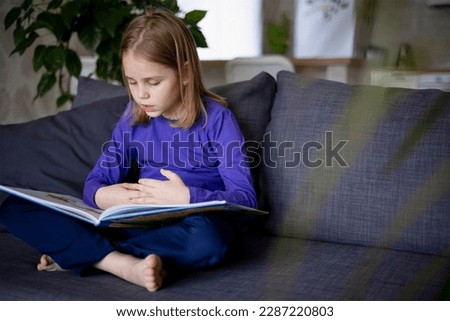 a little blonde girl is reading a big book sitting on the sofa in the living room.Smile and interest in reading.Self-education of children, reading literature at home, interesting leisure