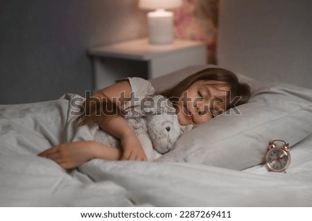 Cute funny cheerful little girl with brown hair sleeping in the white bed with favorite fluffy toy bunny and near lying clock. Copy space. High quality photo