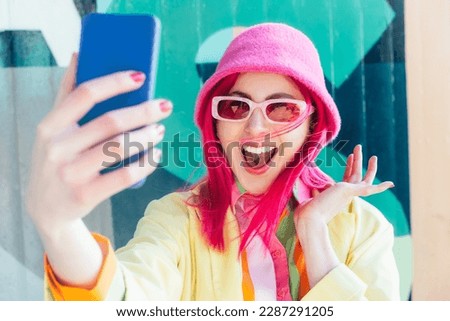Excited pink hair woman screaming while taking a selfie photo outdoors. Emotional hipster fashion woman in bright clothes, pearl pink sunglasses, bucket hat taking selfie photo on the phone camera